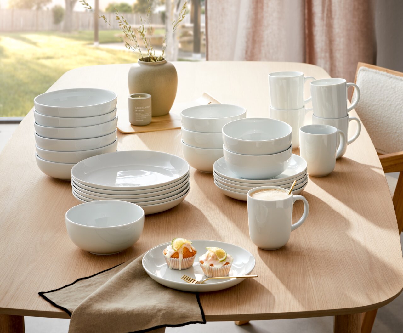 Een 18-delig koffieservies op een tafel, bestaande uit witte borden, schalen en kopjes, in een lichte, moderne eetkamer, zijaanzicht.
