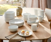 Een 18-delig koffieservies op een tafel, bestaande uit witte borden, schalen en kopjes, in een lichte, moderne eetkamer, zijaanzicht.