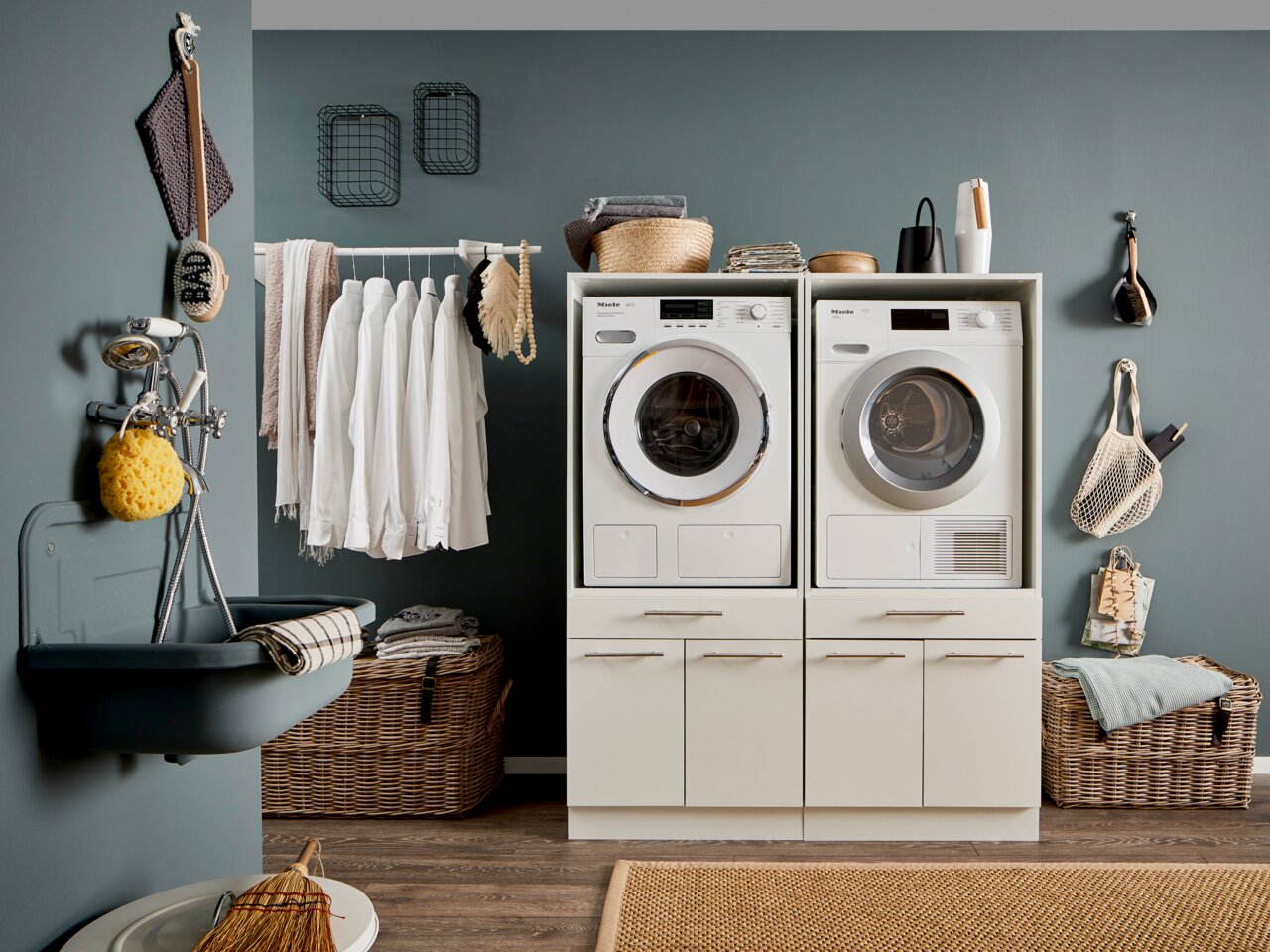 Frontale Ansicht eines modernen Hauswirtschaftsraums mit Waschmaschine und Trockner in einem weißen Schrank, umgeben von Körben und Hängeregalen.