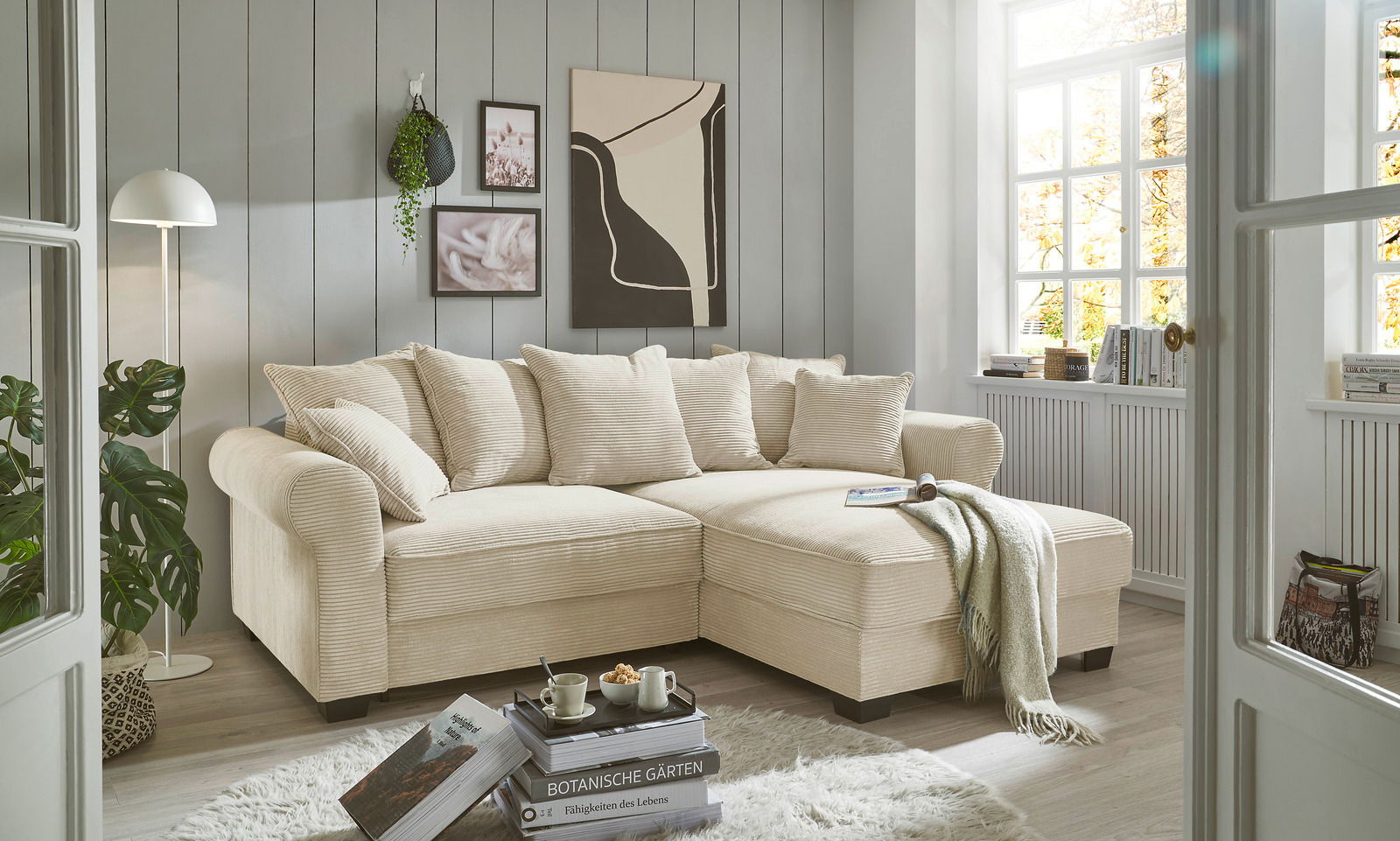 Gemütliche beige Rundecke in einem hellen Wohnzimmer mit großen Fenstern, dekoriert mit Kissen und einer Decke, aus der Perspektive von vorne links.