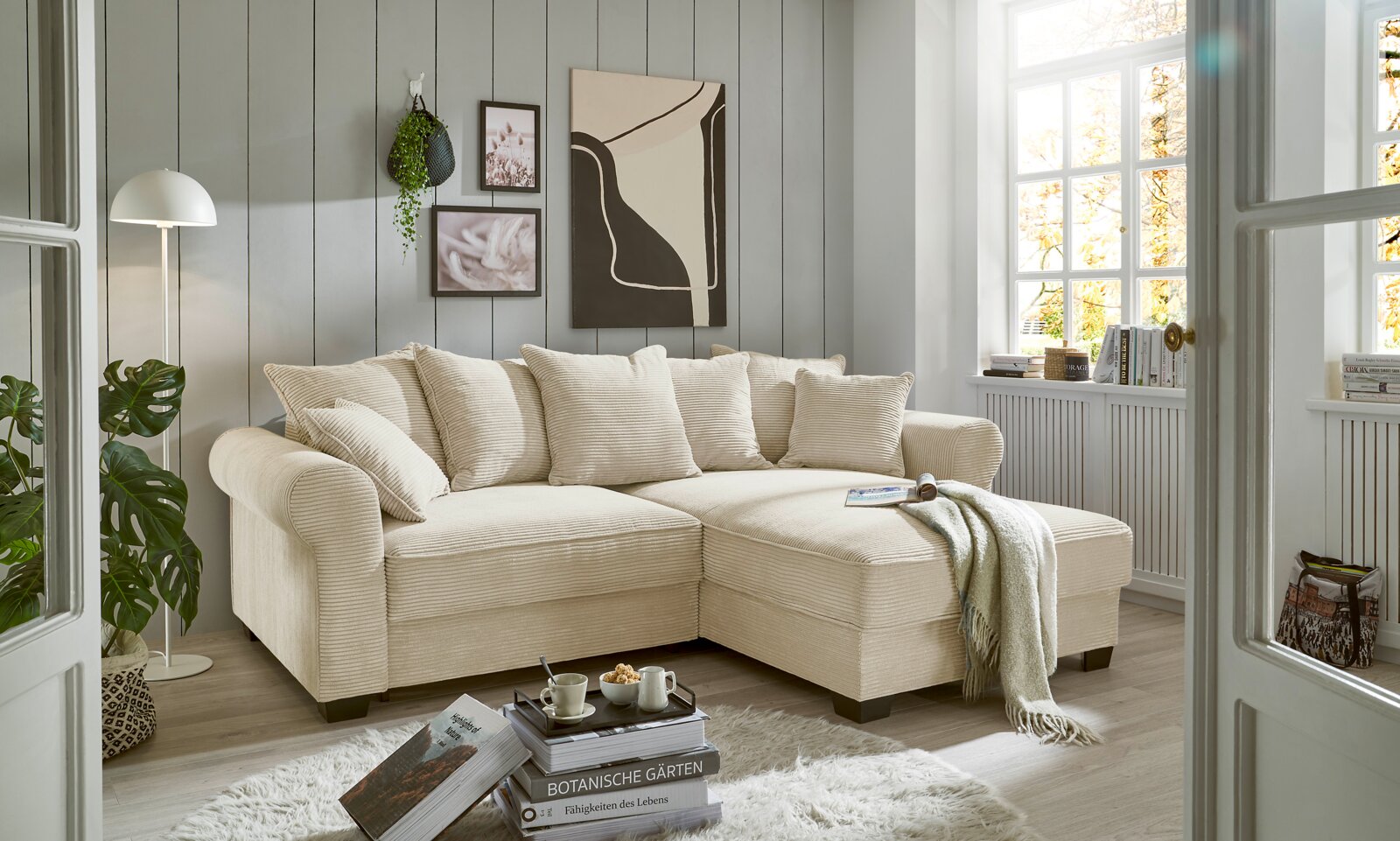 Hom´in Hoekbank MAUREEN Gemütliche beige Rundecke in einem hellen Wohnzimmer mit großen Fenstern, dekoriert mit Kissen und einer Decke, aus der Perspektive von vorne links.