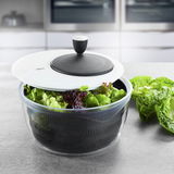Salatschleuder ROTARE mit transparentem Behälter und schwarzem Sieb, gefüllt mit frischem Salat, auf einer Küchenarbeitsplatte aus seitlicher Perspektive.
