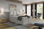 Lichtgrijs gestoffeerd boxspringbed met hoog, verticaal gecapitonneerd hoofdbord, schuin van voren gezien in een moderne slaapkamer.