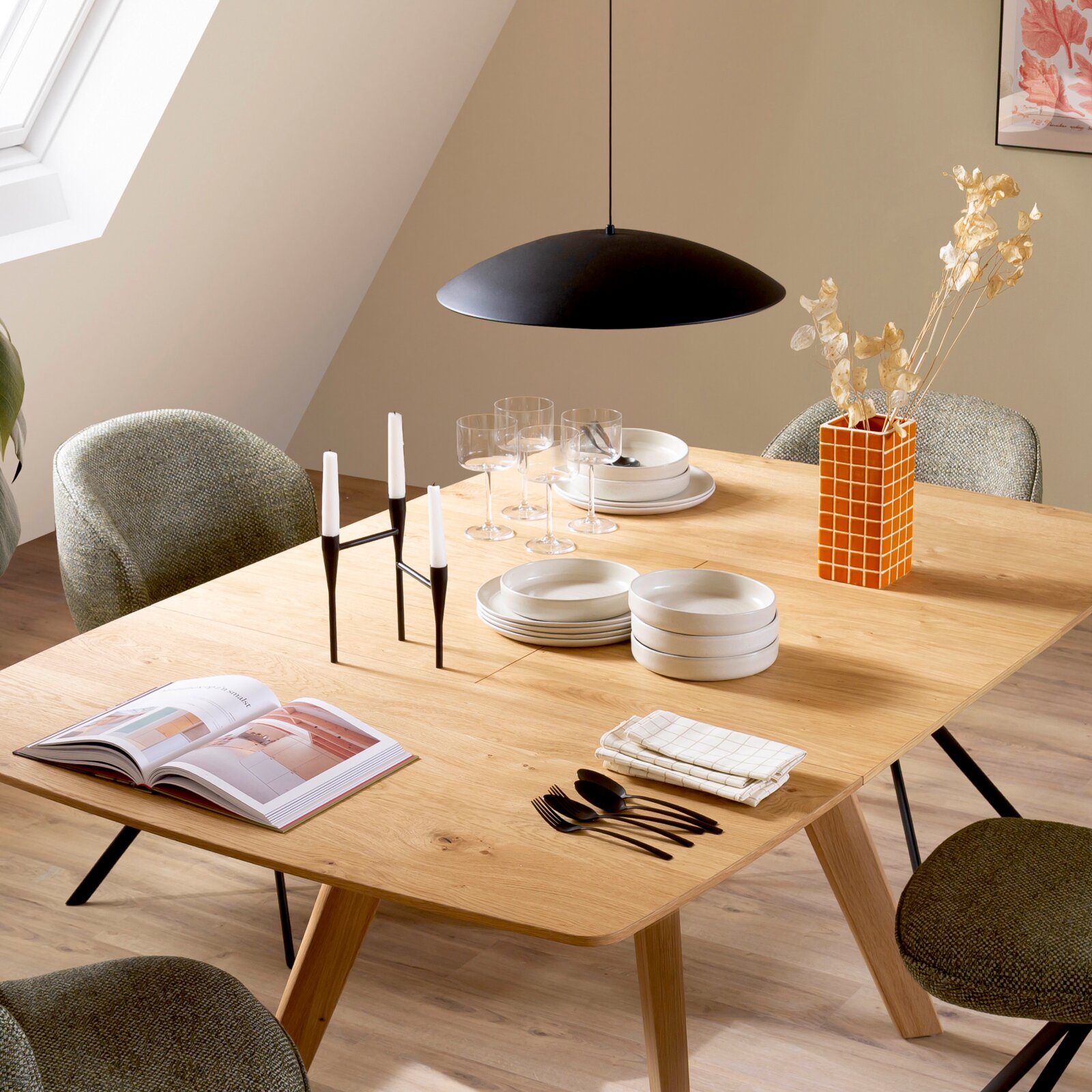 Modern gedeckter Esstisch aus hellem Holz mit schwarzen Metallbeinen, darauf Geschirr, Gläser, Kerzenständer und orangefarbene Vase, darunter schwarze Pendelleuchte; Perspektive: leicht erhöhte Frontansicht schräg von links.