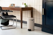 Beiger Treteimer in einem modernen Raum, neben einem Holztisch und einem schwarzen Stuhl, aus seitlicher Perspektive.