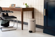 Een beige pedaalemmer in een moderne ruimte, naast een houten tafel en een zwarte stoel, gezien vanuit de zijkant.