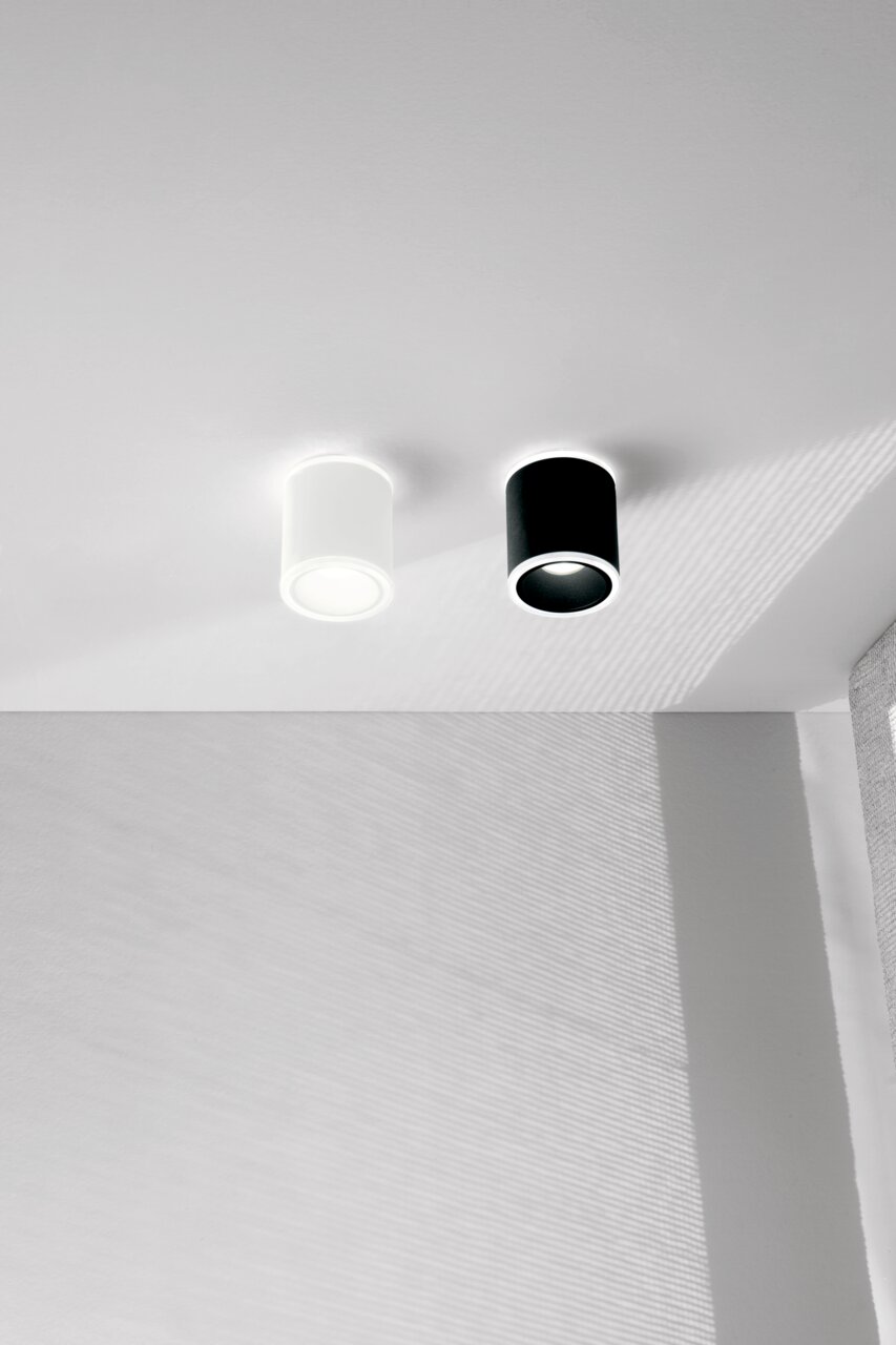 Zwei zylindrische LED-Aufbauspots, einer in Weiß und einer in Schwarz, an einer Decke montiert, seitliche Perspektive.