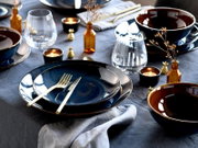 Stilvoll gedeckter Esstisch mit dunkelblau glasierten Tellern und Schalen, goldfarbenem Besteck, klaren Trinkgläsern, Teelichtern und kleinen bernsteinfarbenen Vasen auf grauer Tischdecke, fotografiert in schräger Draufsicht.