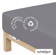 Graues Spannbettlaken aus Bio-Baumwolle und Elasthan, auf einer Matratze gespannt, mit Hinweis auf 100% recycelte Verpackung, seitliche Perspektive.