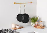 Zwei schwarze 3-PLY Pfannen, aufgehängt an einer goldenen Stange in einer modernen Küche, seitliche Perspektive.