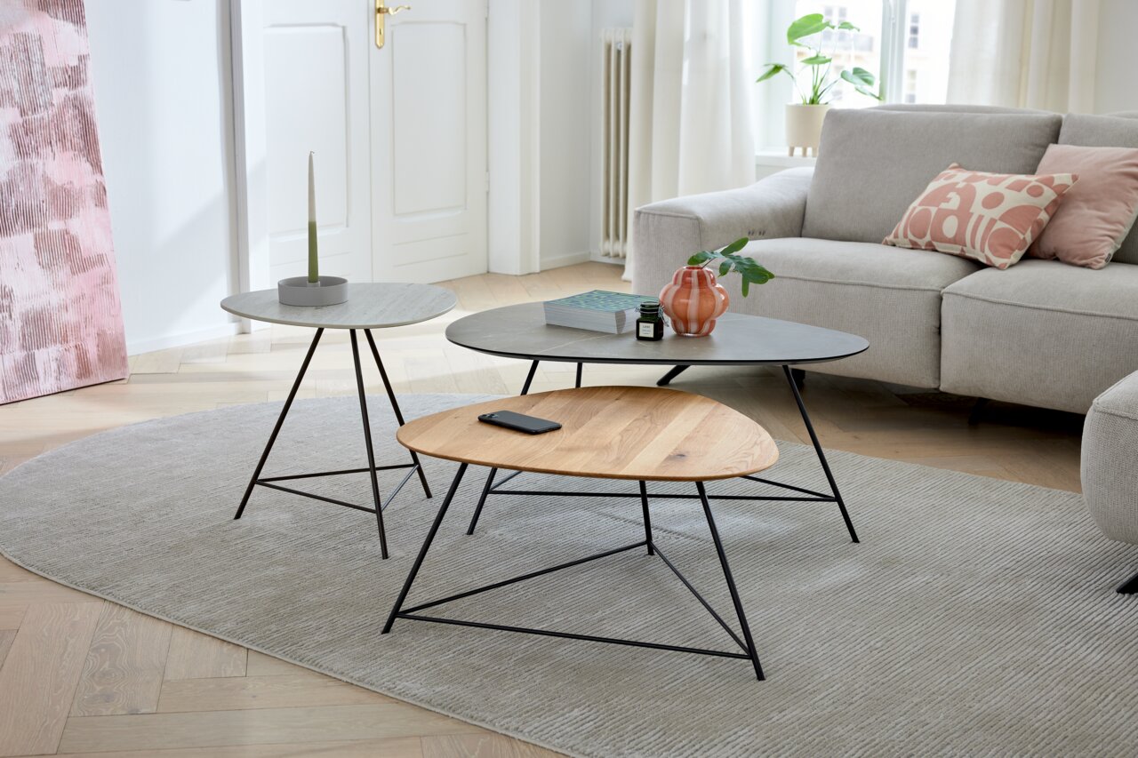 Musterring Couchtisch MR NARA Drei moderne Couchtische in einem Wohnzimmer, mit unterschiedlichen Tischplatten aus Holz und Stein, auf einem Teppich vor einem Sofa. Perspektive von vorne.