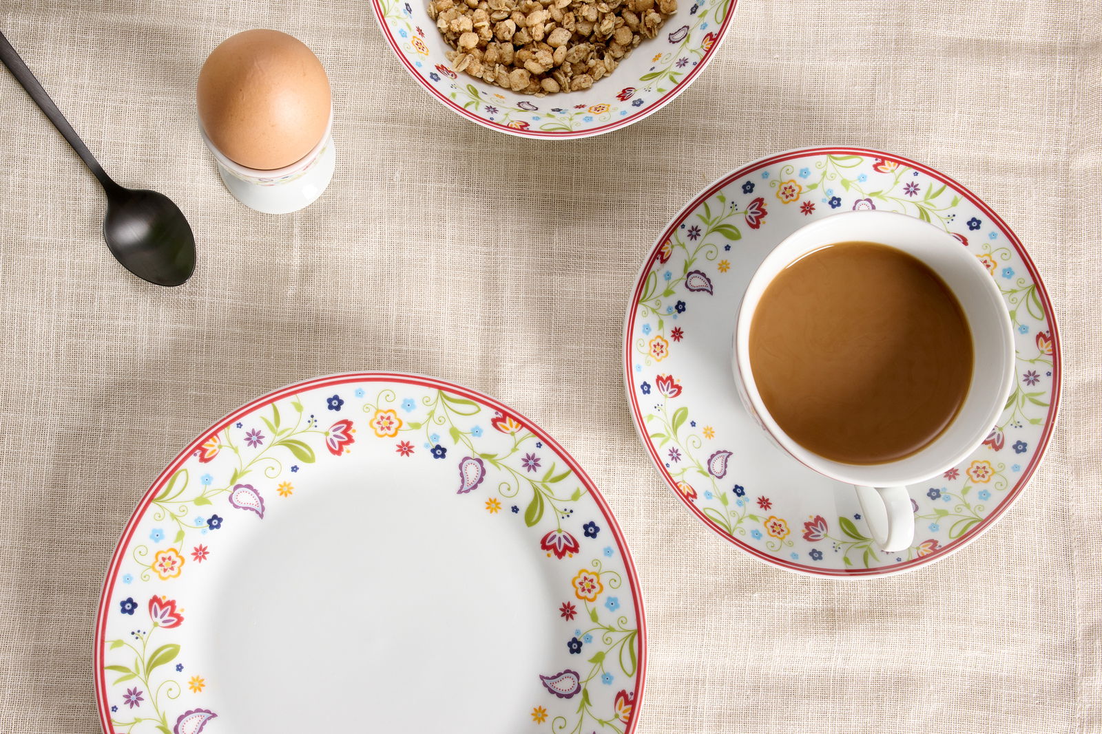 Vogelperspektive auf ein Frühstücksset mit floral verzierten Tellern, einer Tasse Kaffee, einem Ei im Eierbecher und einer Schale Müsli auf einem beigen Tischtuch.