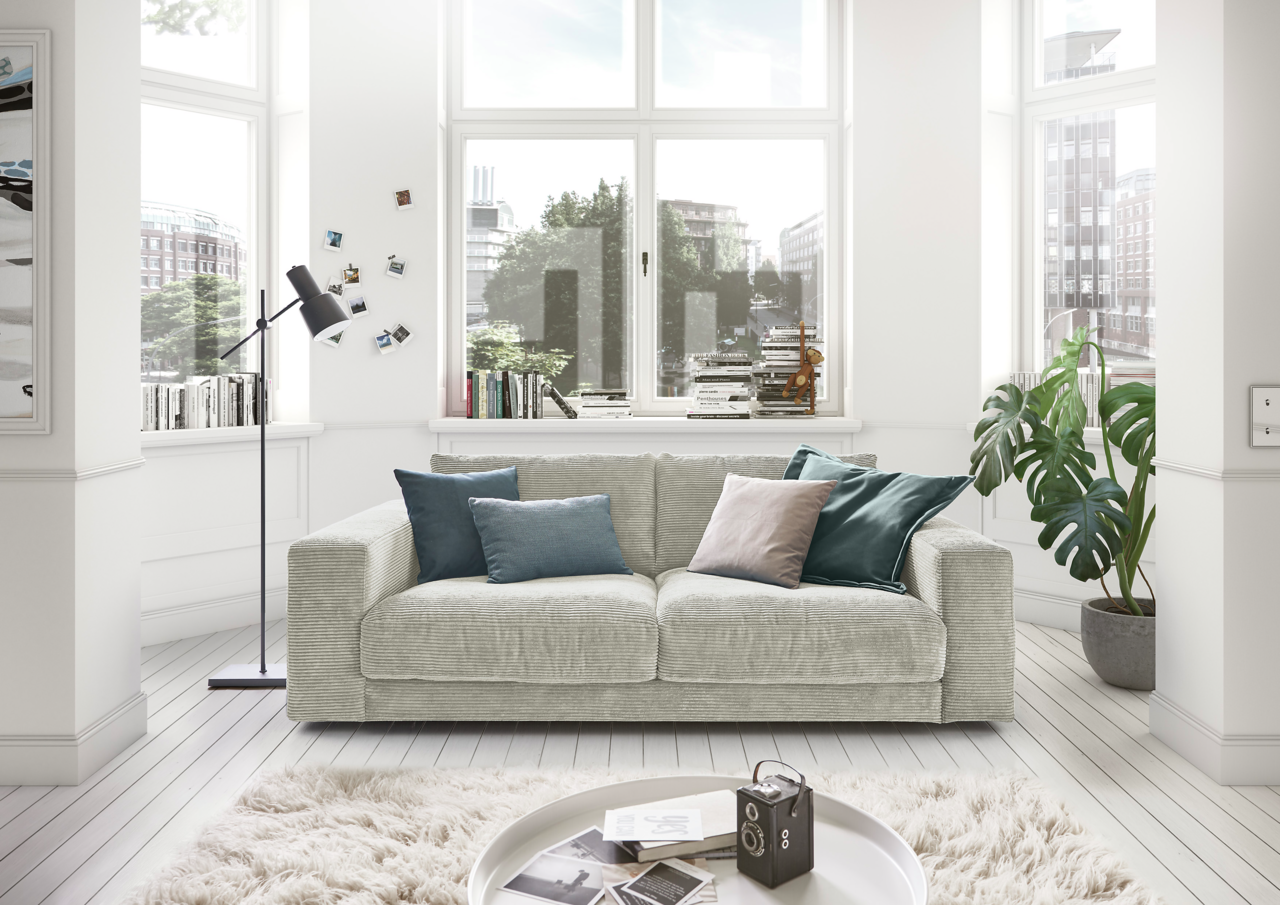 Lounge Handmade Sofa 2-Sitzer LH-SAMBIA Gemütliches 2-Sitzer-Sofa in einem hellen Wohnzimmer mit großen Fenstern, dekoriert mit Kissen in verschiedenen Farben, aus der Frontperspektive.