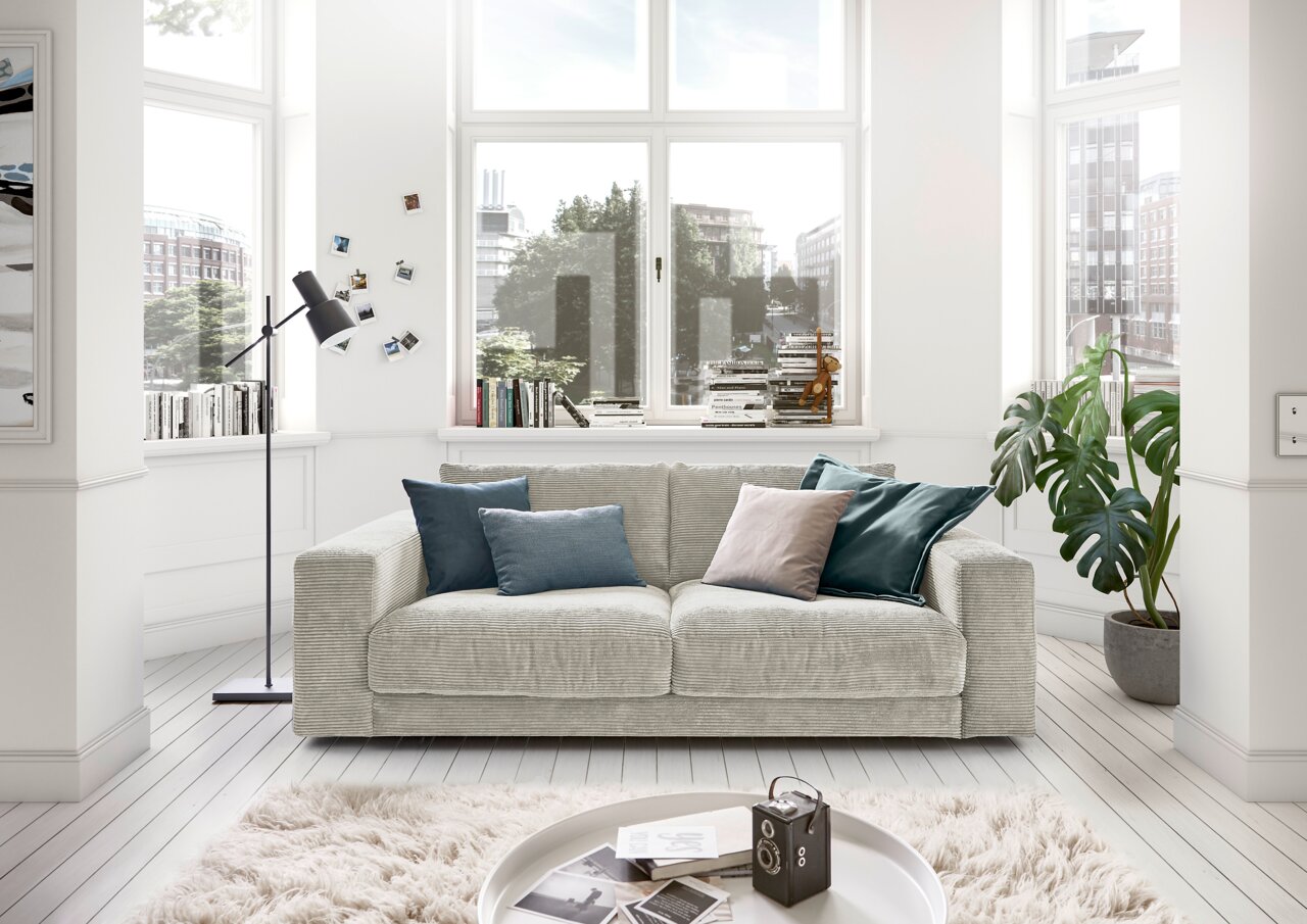 Gemütliches 2-Sitzer-Sofa in einem hellen Wohnzimmer mit großen Fenstern, dekoriert mit Kissen in verschiedenen Farben, aus der Frontperspektive.