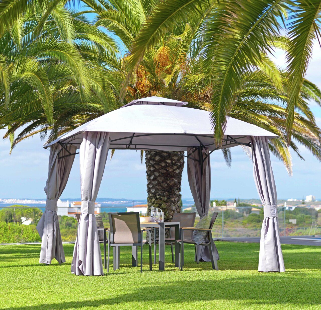 Pavillon im Garten mit grauen Vorhängen, umgeben von Palmen, aus seitlicher Perspektive.