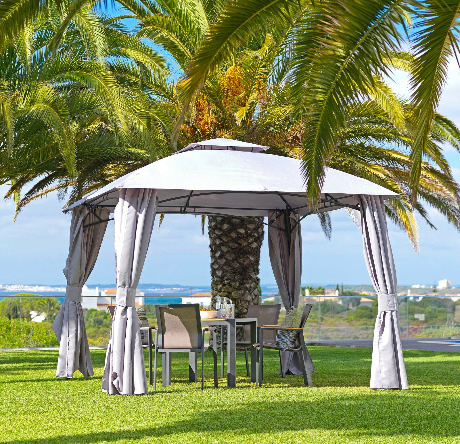 Pavillon im Garten mit grauen Vorhängen, umgeben von Palmen, aus seitlicher Perspektive.