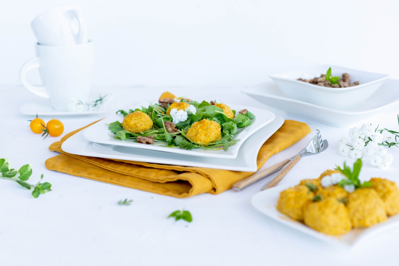 Schräg von vorn fotografierter Teller mit frischem Blattsalat und goldgelben Bällchen auf weißem Geschirr, dekoriert auf heller Tischdecke mit Besteck und gelbem Tuch.