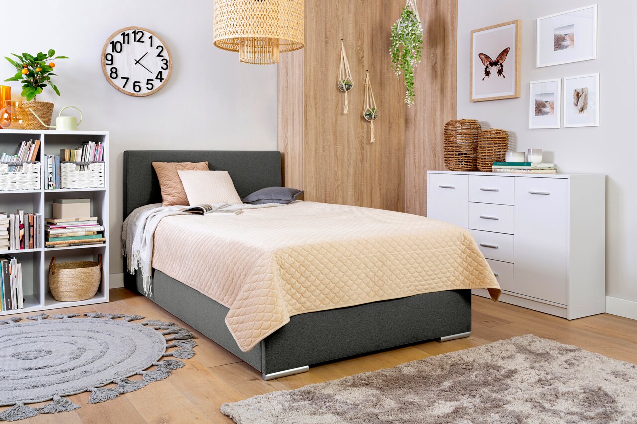 Gemütliches Schlafzimmer mit einem Boxbett in Grau, dekoriert mit Kissen und einer beigen Steppdecke, umgeben von modernen Möbeln und Pflanzen. Perspektive von schräg vorne.
