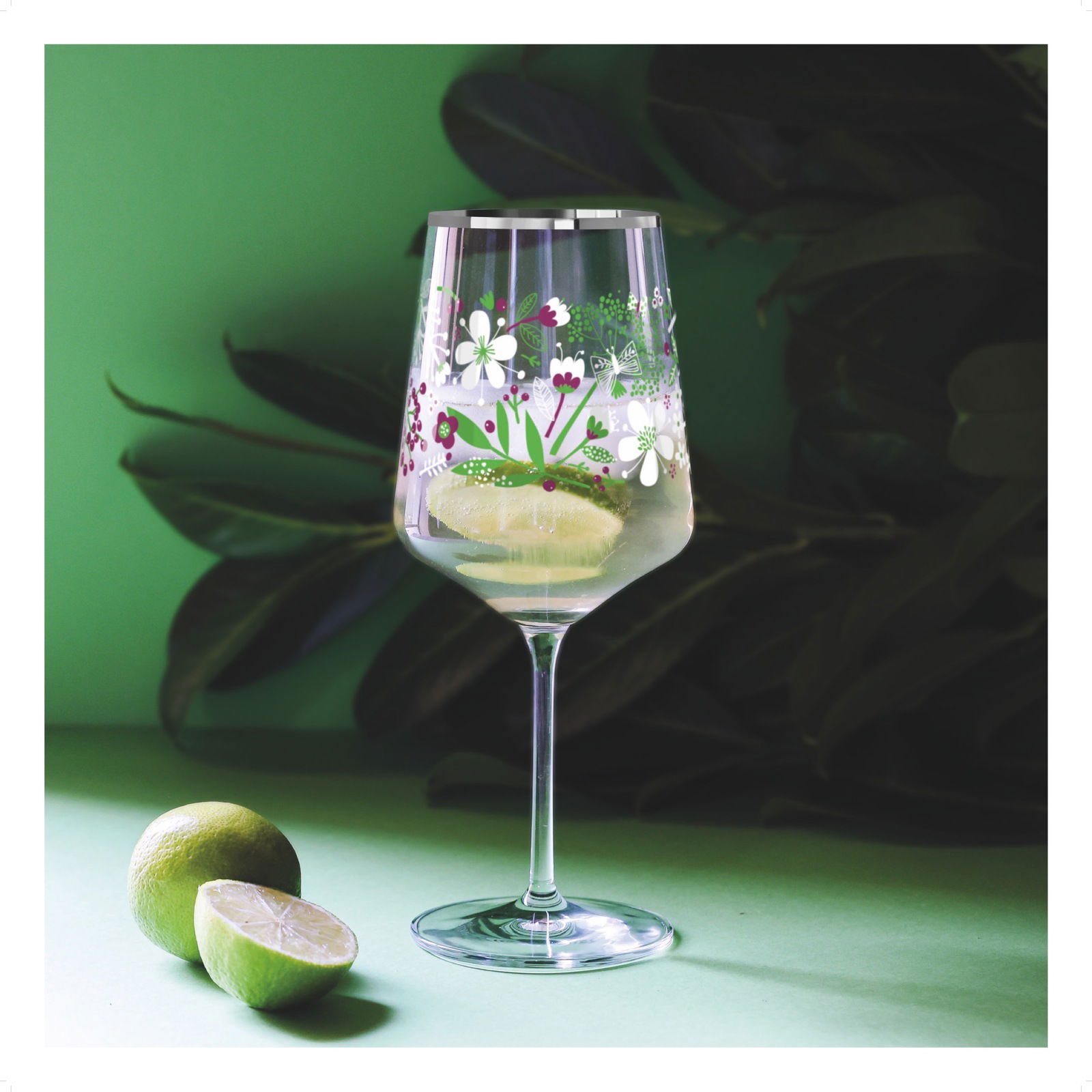 Weinglas mit floralen Mustern, gefüllt mit einem Getränk und Limettenscheiben, auf einem grünen Hintergrund mit Blättern, seitliche Perspektive