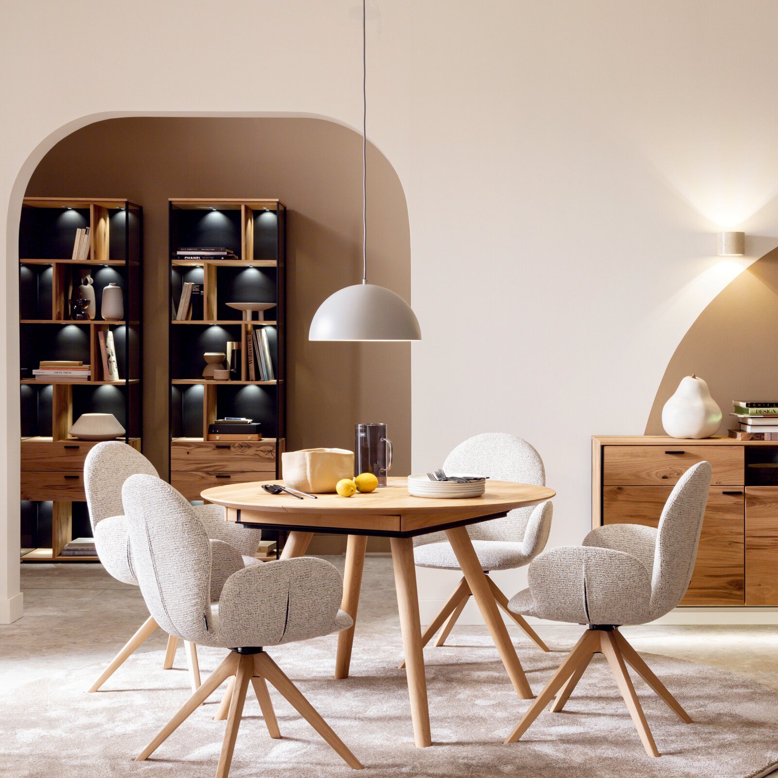 Modernes Esszimmer-Set mit rundem Holztisch und vier gepolsterten, hellgrauen Stühlen, darüber eine weiße Pendelleuchte; frontal in leichter Seitenperspektive aufgenommen.
