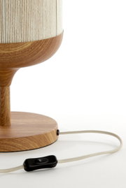 Detailfoto van een tafellamp met houten voet en houten sokkel, crèmekleurige lampenkap met structuur en stoffen snoer met zwarte tussenschakelaar, close-up van de zijkant