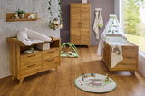 Babyzimmer mit Holz-Babybett, Wickelkommode und Schrank in warmer Holzoptik, von der Seite betrachtet. Helle, freundliche Atmosphäre mit Spielmatte und Dekorationen.