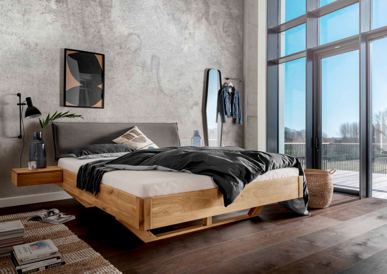 Massivholzbettgestell aus hellem Holz mit grauem Kopfteil, seitlicher Perspektive, in modernem Schlafzimmer mit großen Fenstern.