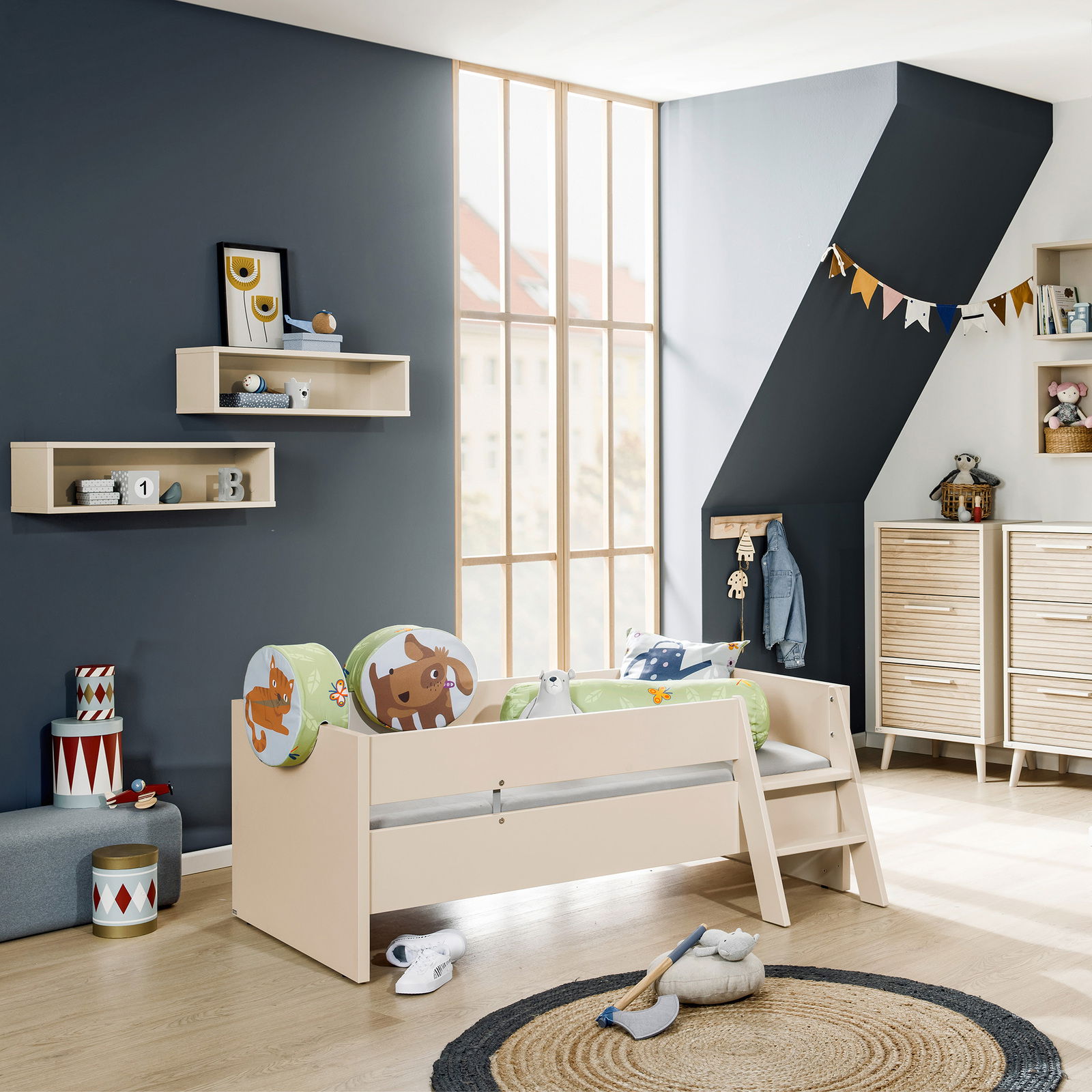 Kinderzimmer mit einem beige-farbenen Kinderbett, das mit bunten Tierkissen dekoriert ist. Das Bett hat eine kleine Leiter und steht in einem hellen Raum mit großen Fenstern. Perspektive: seitliche Ansicht des Bettes.