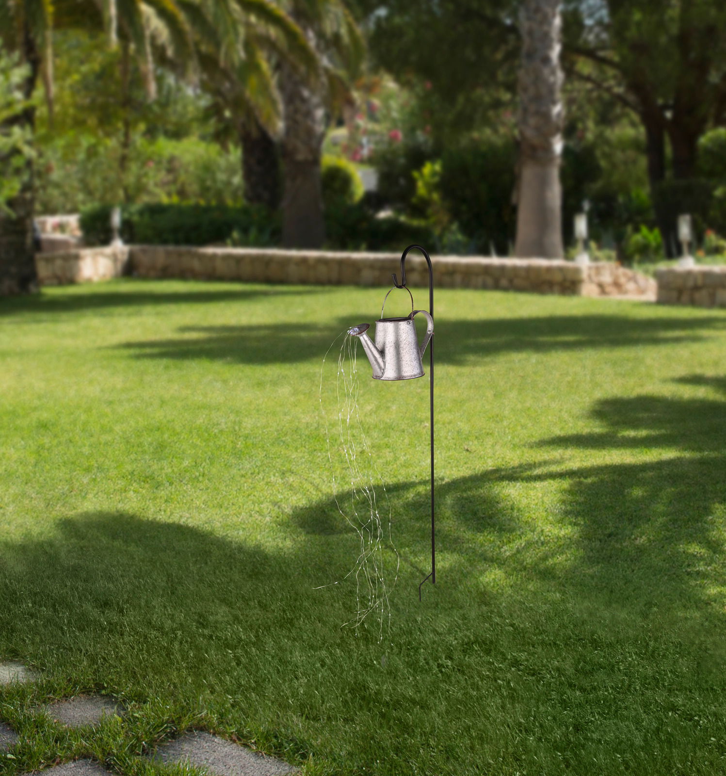 Gartenansicht mit einer Solarleuchte in Form einer Gießkanne, die an einem Metallstab hängt und Lichterketten wie Wasserstrahlen ausstrahlt.