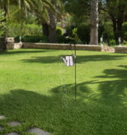 Gartenansicht mit einer Solarleuchte in Form einer Gießkanne, die an einem Metallstab hängt und Lichterketten wie Wasserstrahlen ausstrahlt.