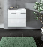 Modernes weißes, wandhängendes Badezimmer-Waschbecken mit Unterschrank und zwei Türen mit Metallgriffen vor grauer Fliesenwand, frontal aus Augenhöhe aufgenommen; unten grüner Badteppich und rechts eine Topfpflanze