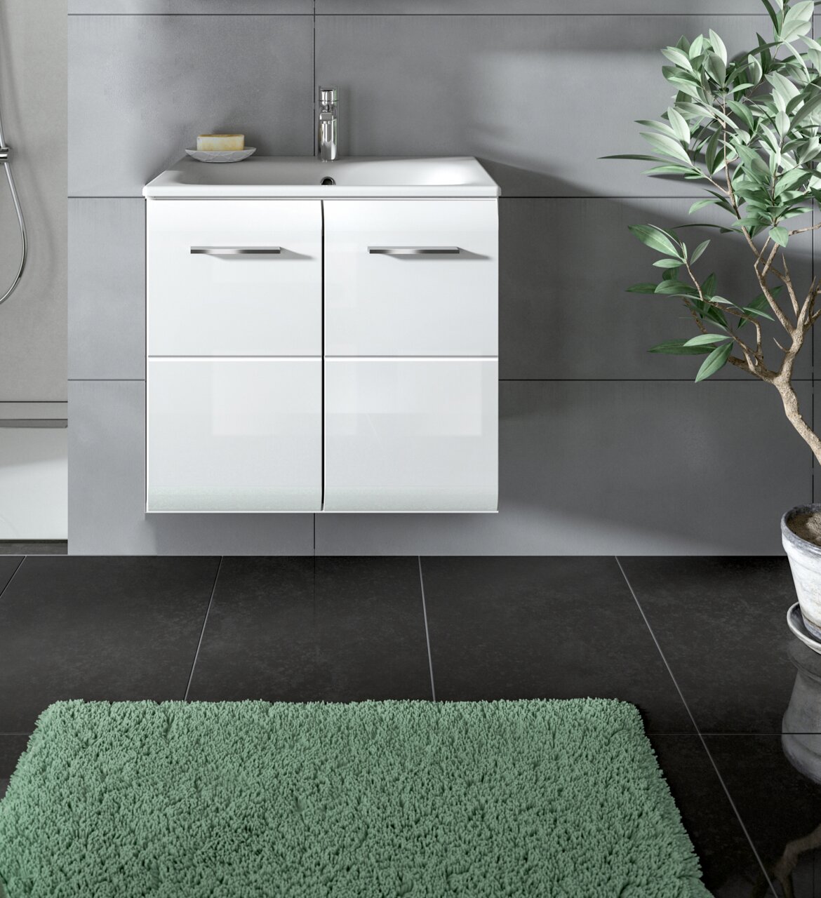 Modernes weißes, wandhängendes Badezimmer-Waschbecken mit Unterschrank und zwei Türen mit Metallgriffen vor grauer Fliesenwand, frontal aus Augenhöhe aufgenommen; unten grüner Badteppich und rechts eine Topfpflanze