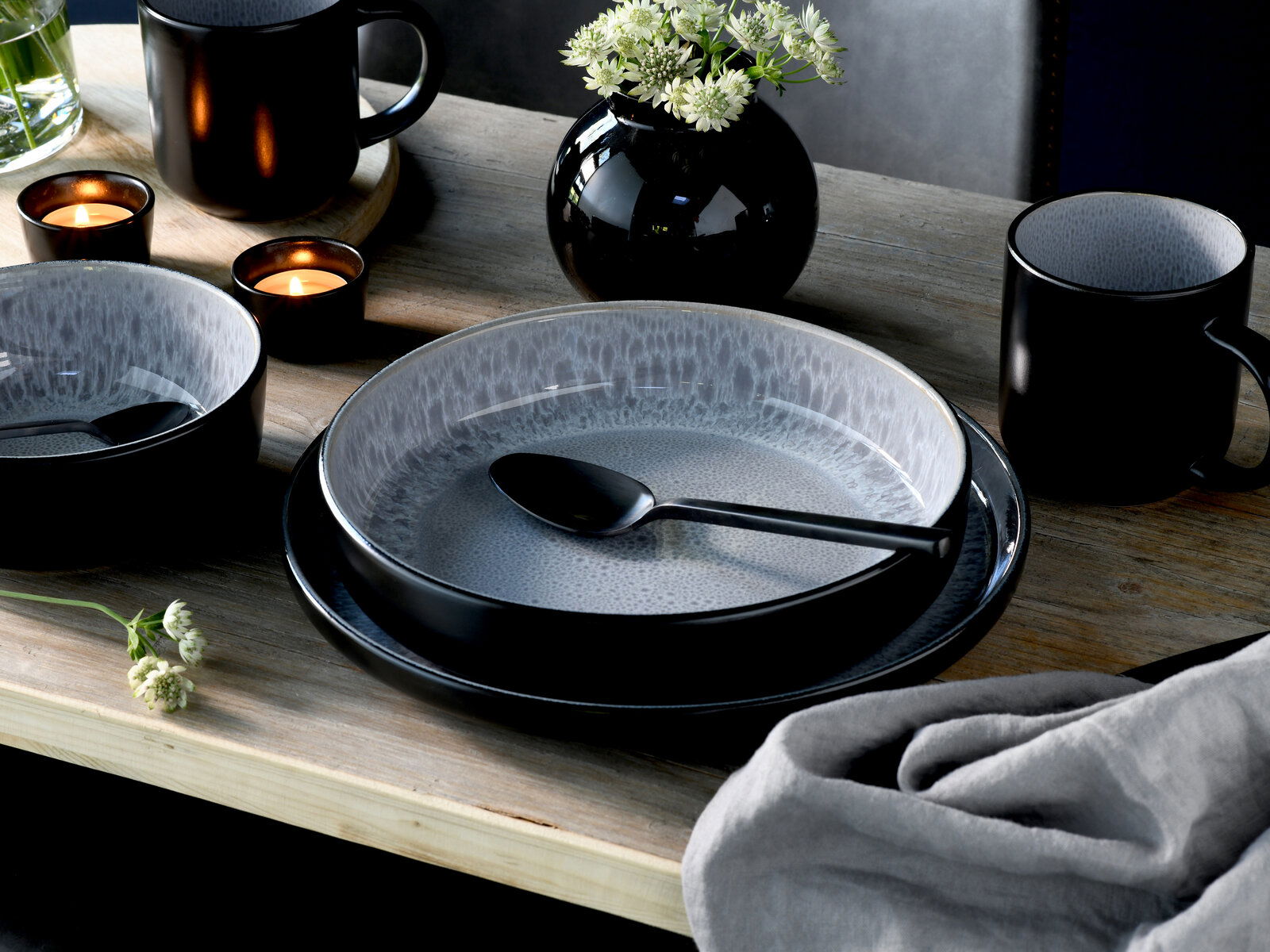 Elegantes Kombiservice 30-tlg. Frost in Schwarz und Grau, bestehend aus Tellern, Schalen und Tassen, auf einem Holztisch mit Kerzen und Blumen, aus der Vogelperspektive fotografiert.