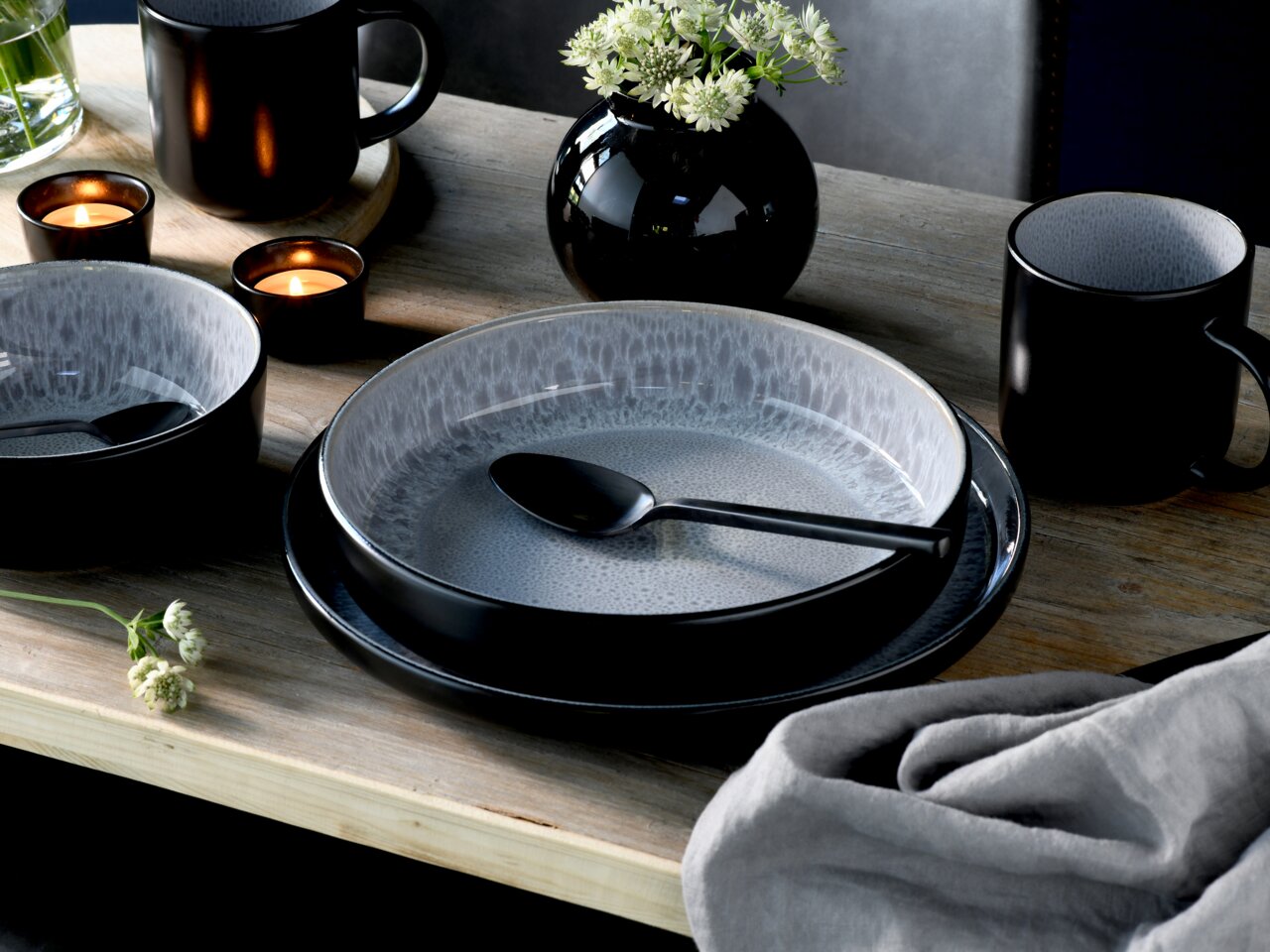 Elegantes Kombiservice 30-tlg. Frost in Schwarz und Grau, bestehend aus Tellern, Schalen und Tassen, auf einem Holztisch mit Kerzen und Blumen, aus der Vogelperspektive fotografiert.