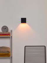 Moderne schwarze Wandleuchte IO an einer weißen Wand, die warmes Licht nach unten abstrahlt. Perspektive: Frontalansicht.