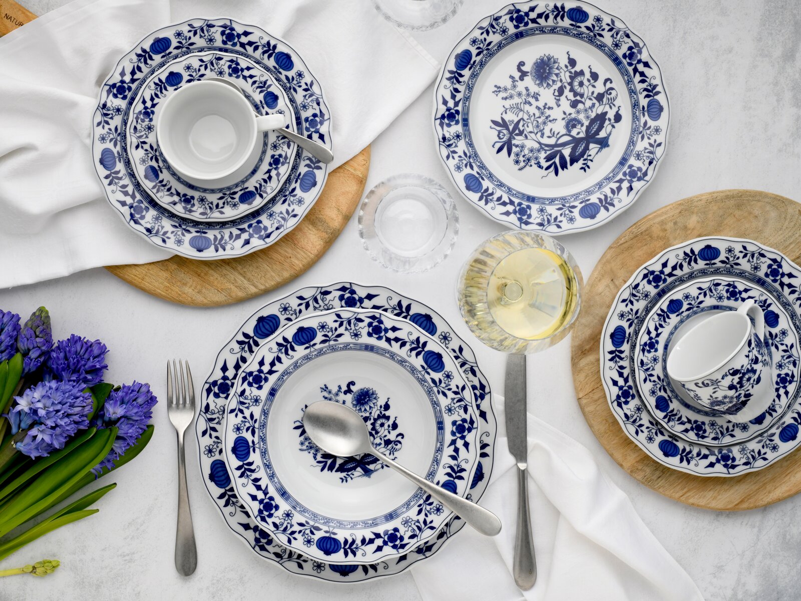 Dunkelblau-weißes Porzellan-Geschirrset mit floralem Dekor, Tellern und Tassen auf weißer Tischdecke mit Besteck, Holzuntersetzern, Glas und Blumen, fotografiert in Draufsicht von oben