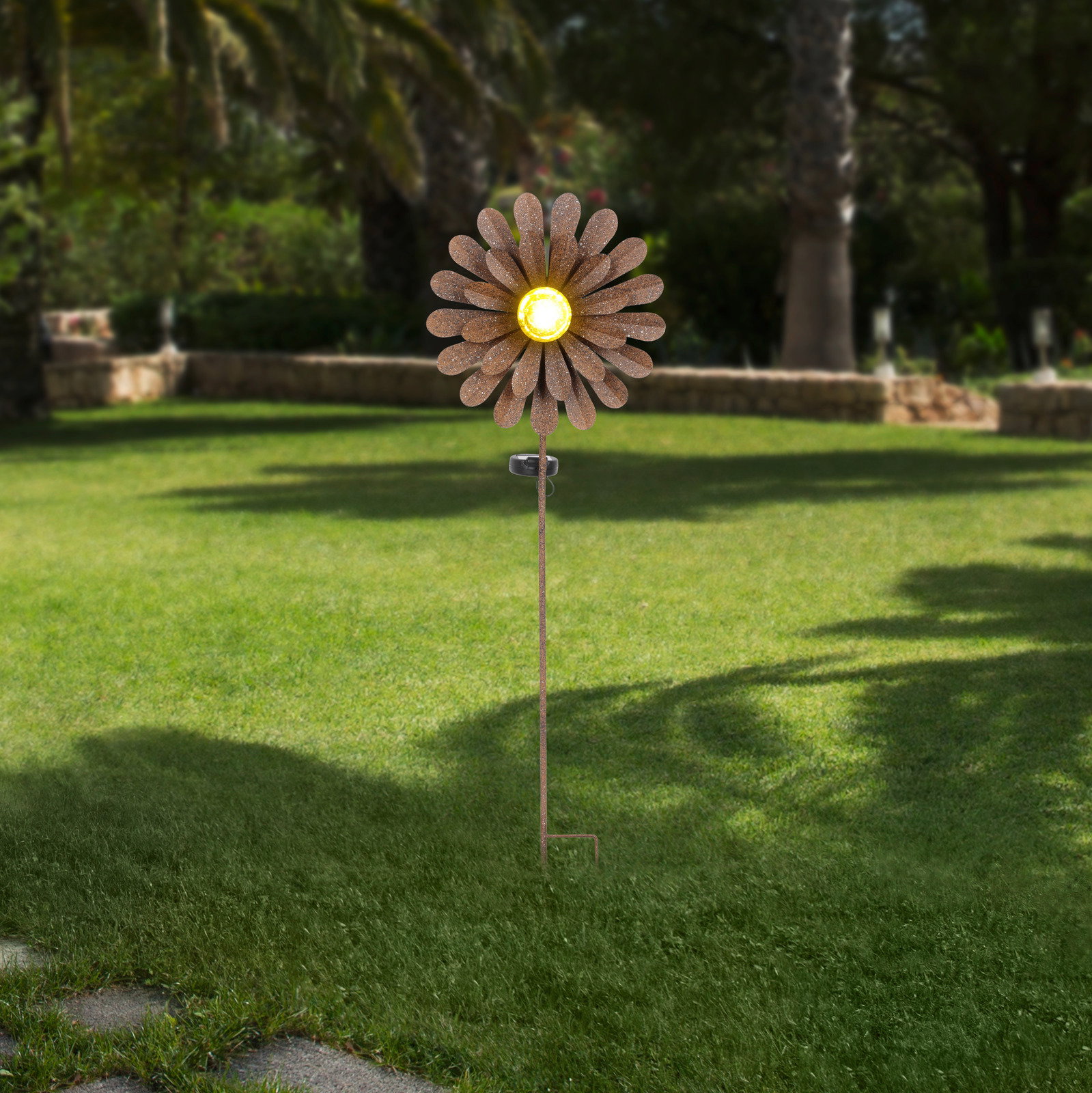 Solarleuchte in Form einer Blume mit gelbem Licht in der Mitte, aufgestellt in einem Garten. Frontalansicht.