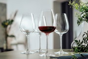 Vier elegante Rotweingläser, eines davon mit Rotwein gefüllt, auf einem Tisch in einem modernen, stilvollen Ambiente. Perspektive: seitlich, leicht von oben.