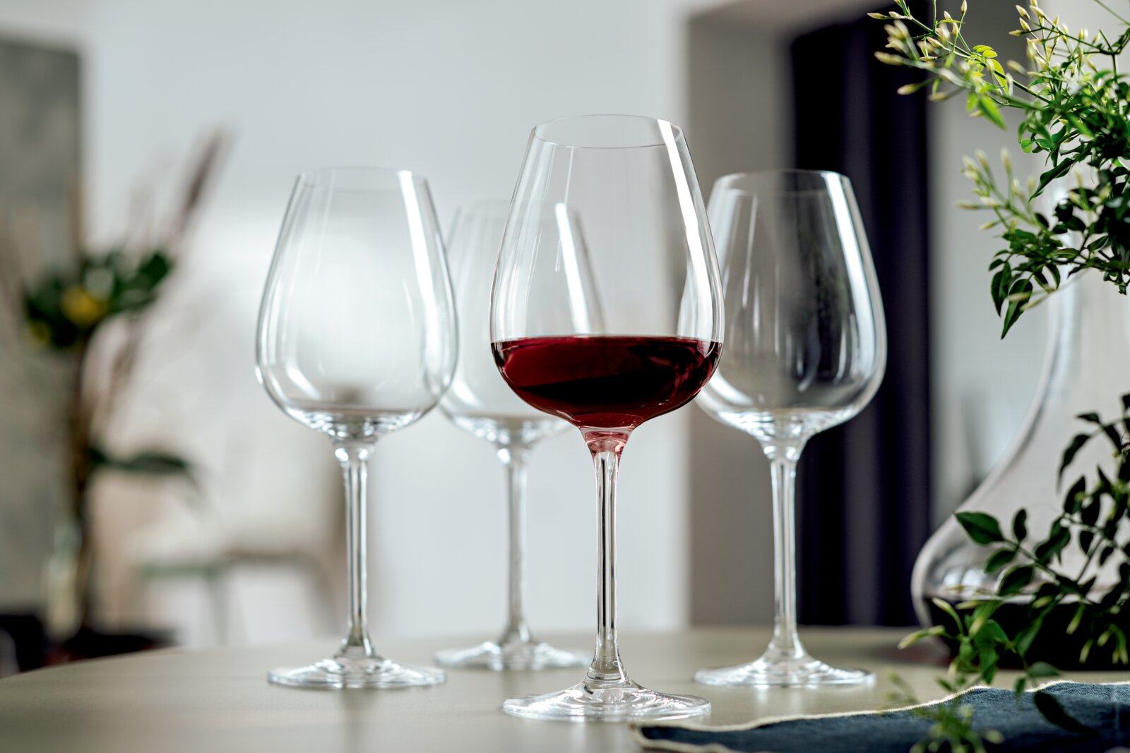 Vier elegante Rotweingläser, eines davon mit Rotwein gefüllt, auf einem Tisch in einem modernen, stilvollen Ambiente. Perspektive: seitlich, leicht von oben.