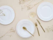 Minimalistisches Tischgedeck in Draufsicht: drei weiße Teller/Schalen auf hellem Untergrund mit goldfarbenem Besteck (Löffel, Gabel, Messer), Weinglas und dekorativen Trockenblumen.
