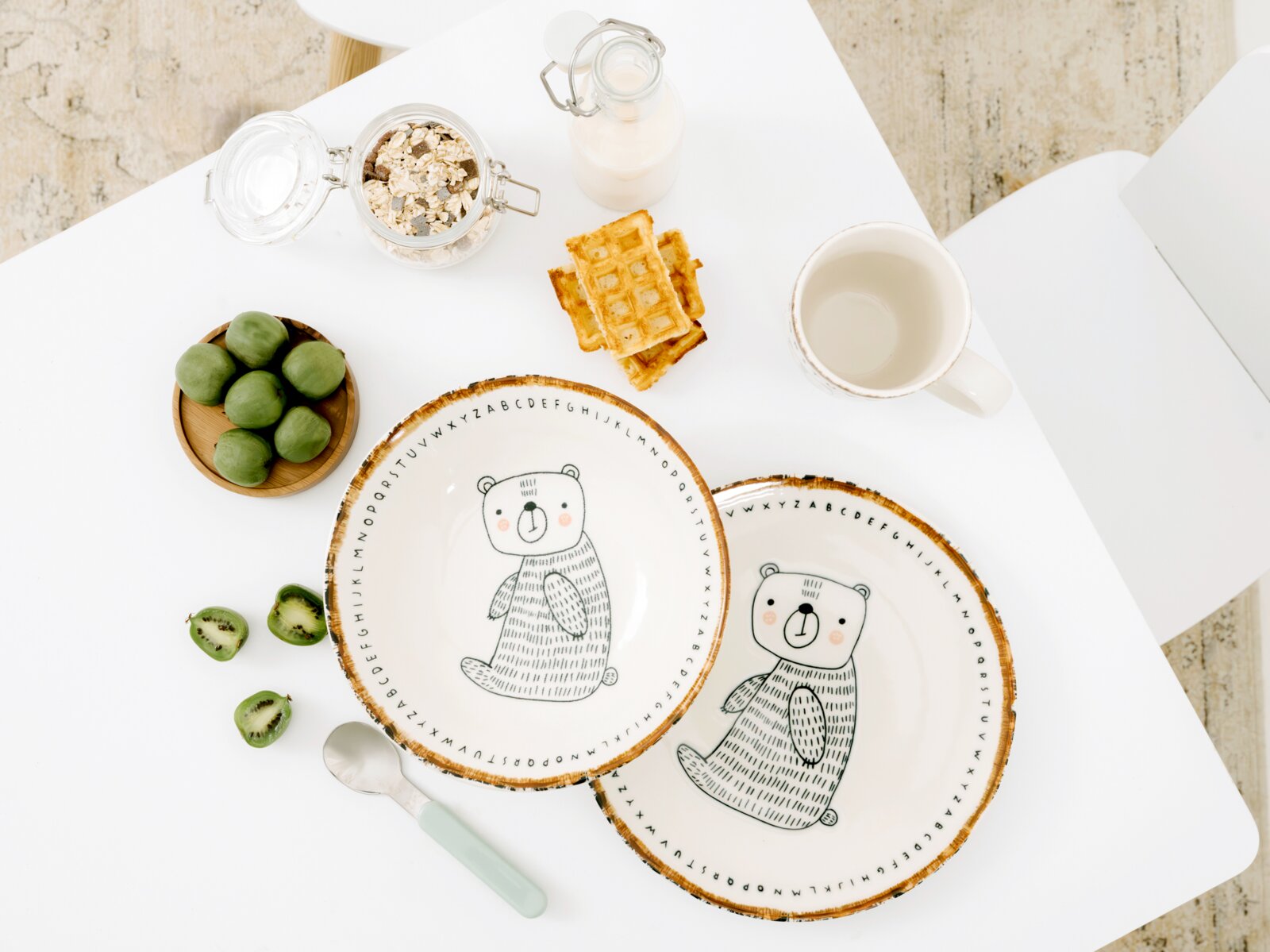 Zwei cremefarbene Kinderteller mit niedlichem Bärenmotiv, Alphabet-Rand und rustikaler brauner Kante, aus der Vogelperspektive auf einem gedeckten Frühstückstisch präsentiert
