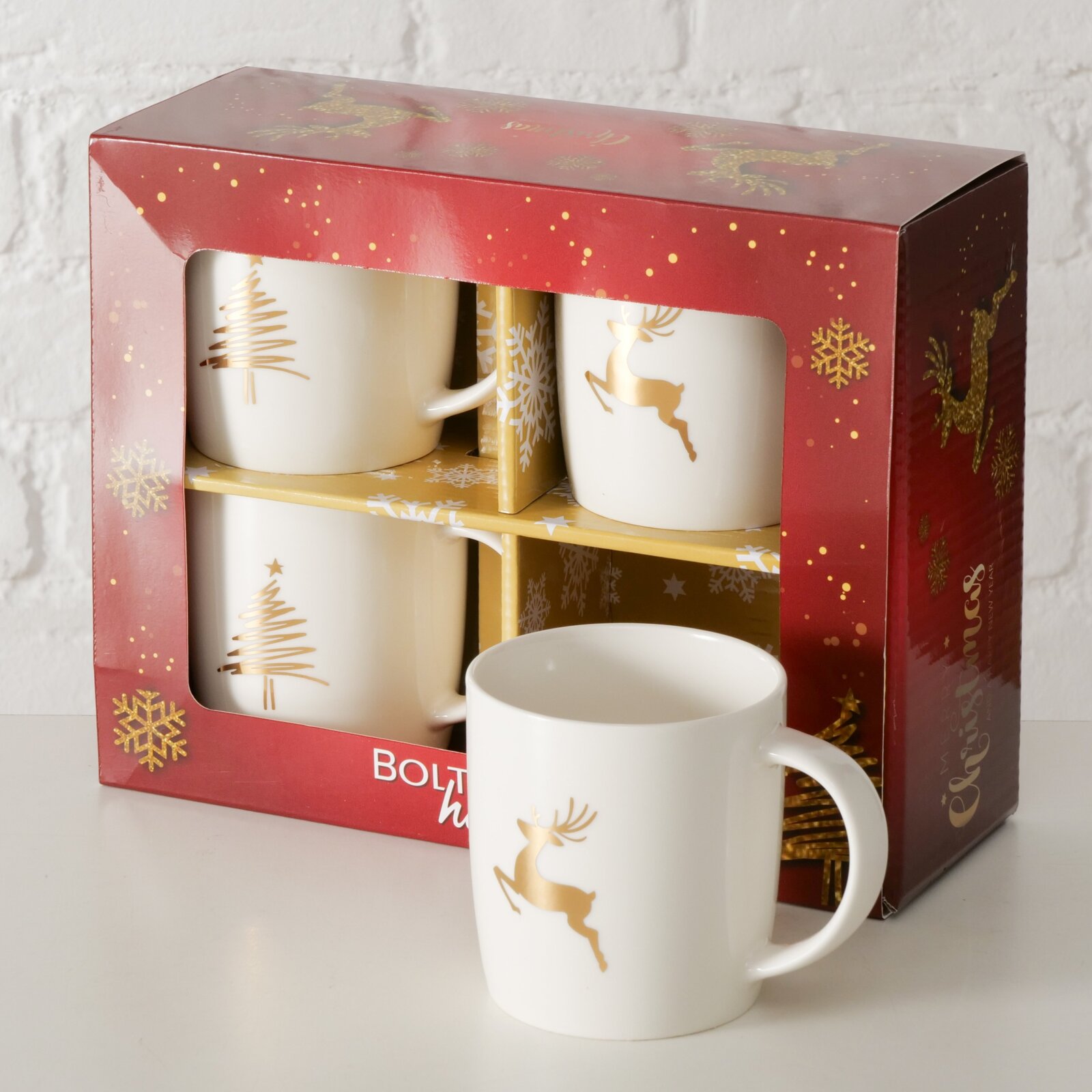 Vier witte bekers met gouden kerstmotieven, waaronder een rendier en een kerstboom, in een feestelijke rode geschenkdoos. Perspectief: licht van bovenaf en van opzij.