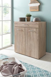 Wäschekommode aus Holz mit zwei Türen und einer Schublade, dekoriert mit Pflanzen und einem Hut, in einem hellen Raum mit Fenster, aus seitlicher Perspektive.