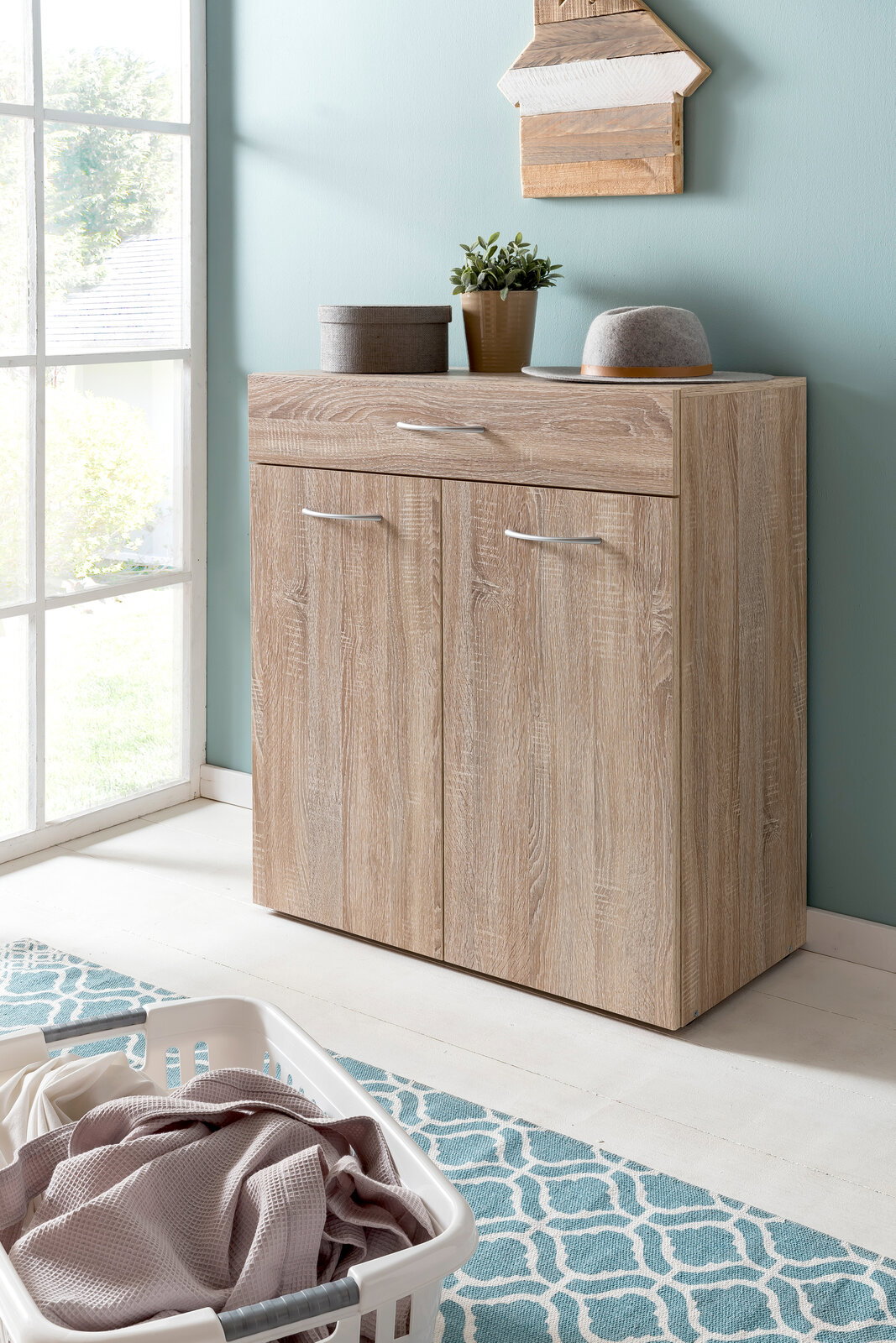 XORA Wäschekommode MULTIRAUMKONZEPT Wäschekommode aus Holz mit zwei Türen und einer Schublade, dekoriert mit Pflanzen und einem Hut, in einem hellen Raum mit Fenster, aus seitlicher Perspektive.