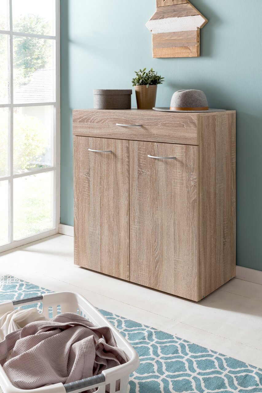 Wäschekommode aus Holz mit zwei Türen und einer Schublade, dekoriert mit Pflanzen und einem Hut, in einem hellen Raum mit Fenster, aus seitlicher Perspektive.