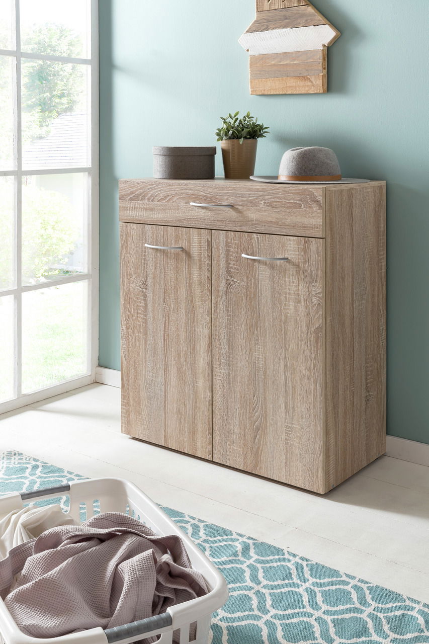 Wäschekommode aus Holz mit zwei Türen und einer Schublade, dekoriert mit Pflanzen und einem Hut, in einem hellen Raum mit Fenster, aus seitlicher Perspektive.
