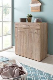 Wäschekommode aus Holz mit zwei Türen und einer Schublade, dekoriert mit Pflanzen und einem Hut, in einem hellen Raum mit Fenster, aus seitlicher Perspektive.