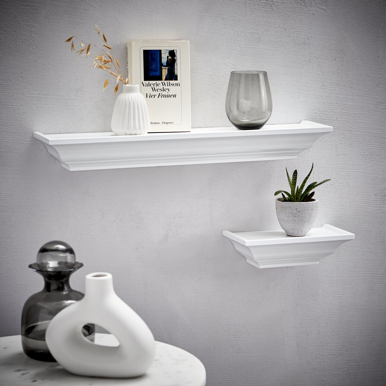 Zwei weiße Wandboards der Marke XORA, Modell VIOLA, an einer grauen Wand. Das größere Board trägt eine Vase, ein Buch und ein Glas, das kleinere eine Pflanze. Perspektive von vorne.