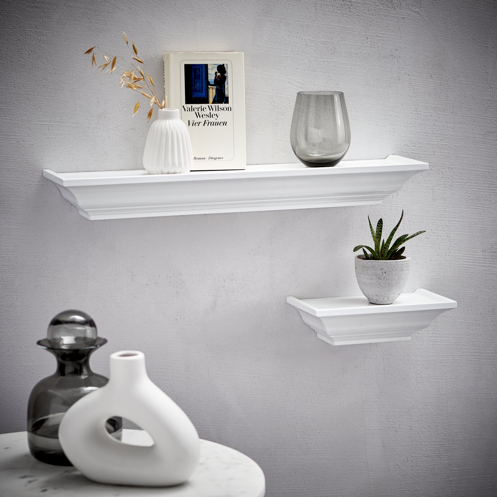 Zwei weiße Wandboards der Marke XORA, Modell VIOLA, an einer grauen Wand. Das größere Board trägt eine Vase, ein Buch und ein Glas, das kleinere eine Pflanze. Perspektive von vorne.