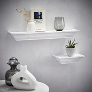 Zwei weiße Wandboards der Marke XORA, Modell VIOLA, an einer grauen Wand. Das größere Board trägt eine Vase, ein Buch und ein Glas, das kleinere eine Pflanze. Perspektive von vorne.